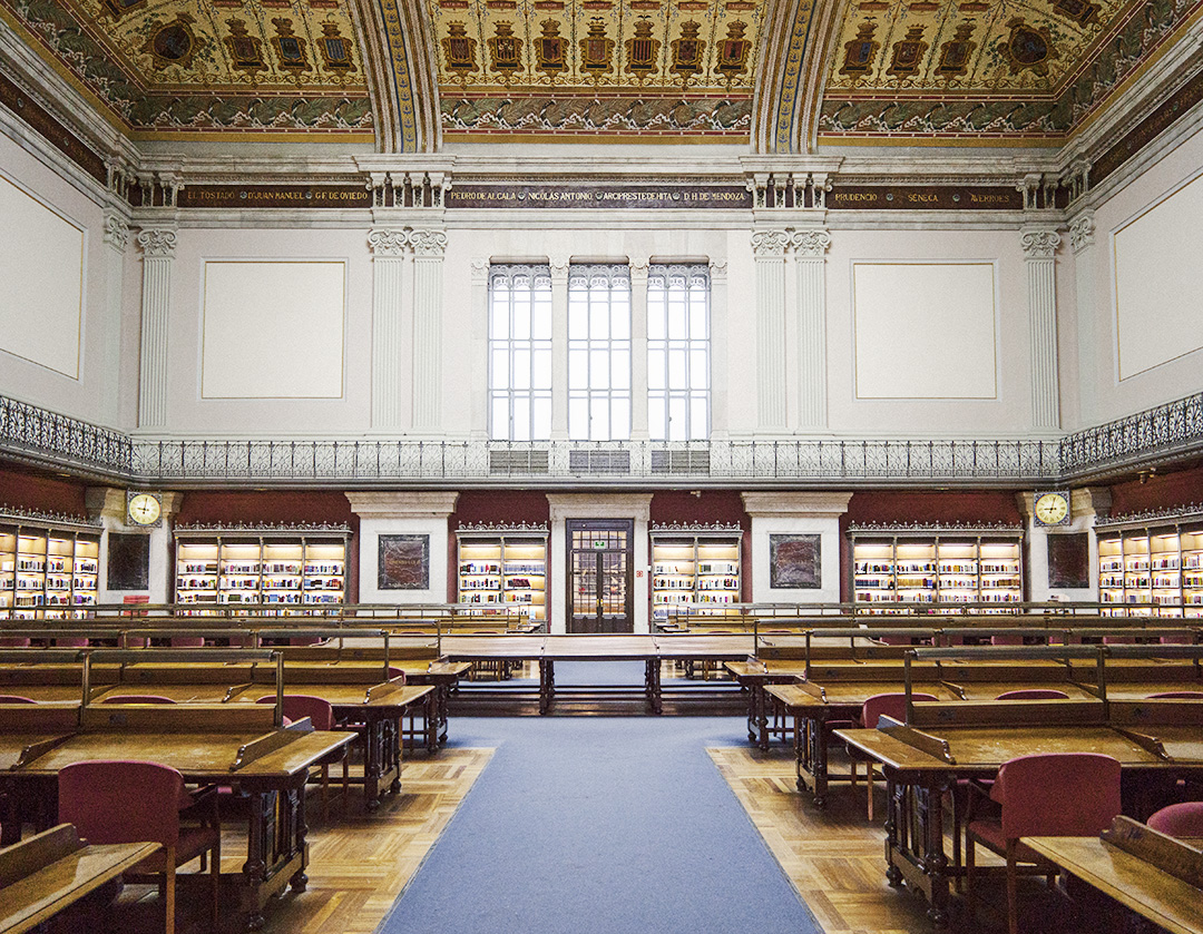 Sala de lectura María Moliner de la Biblioteca Nacional de España, un gran salón con mesas de madera, estanterías iluminadas y altos techos decorados con frescos, vista frontal y simétrica.